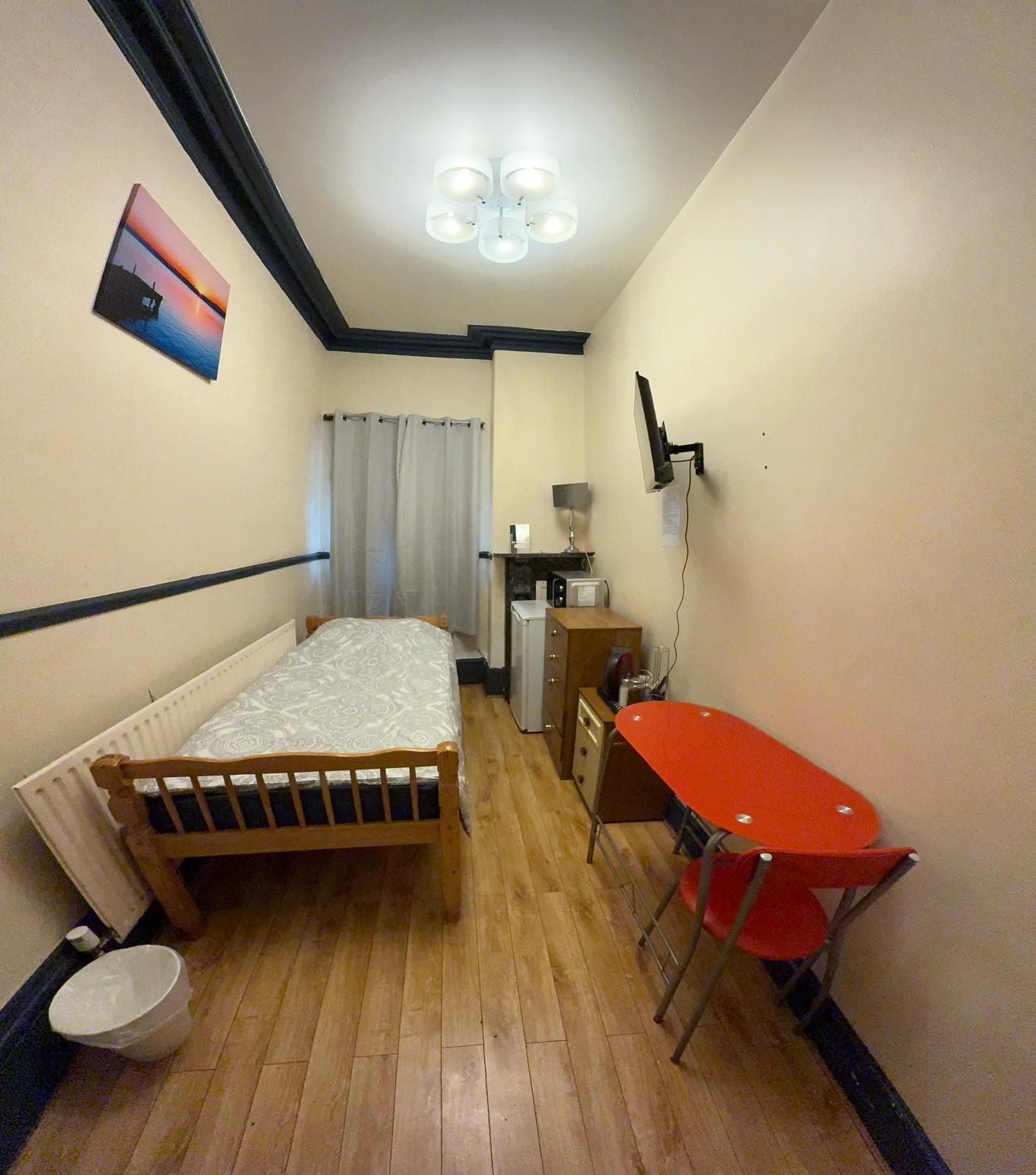A small, narrow room with a bed, a desk, a chair, a radiator, and a wall-mounted TV.