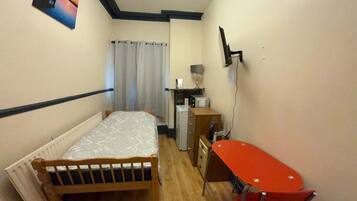 A small, narrow room with a bed, a desk, a chair, a radiator, and a wall-mounted TV.