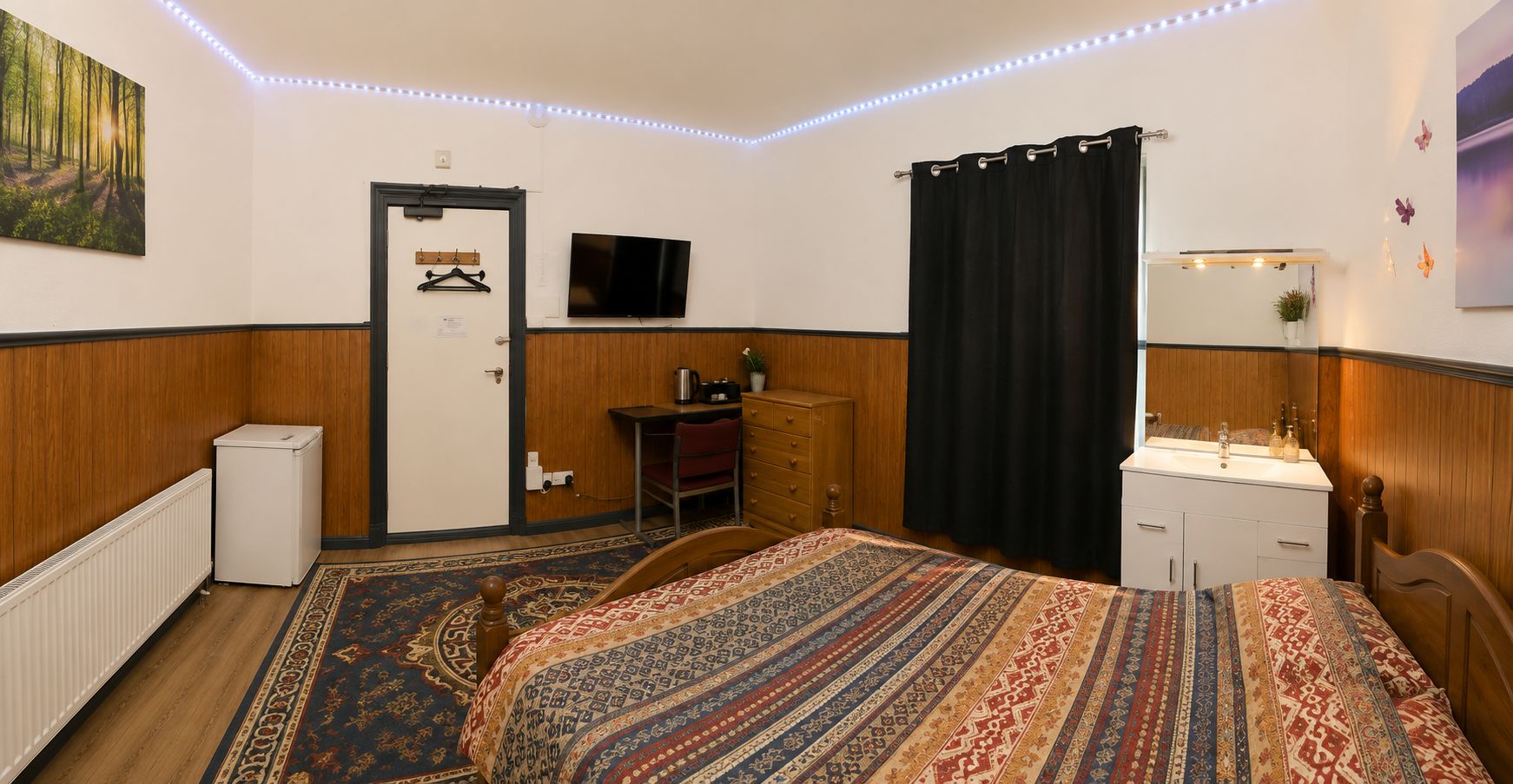 Una habitación de hotel con cama, escritorio, televisor, radiador y ventana con cortinas.