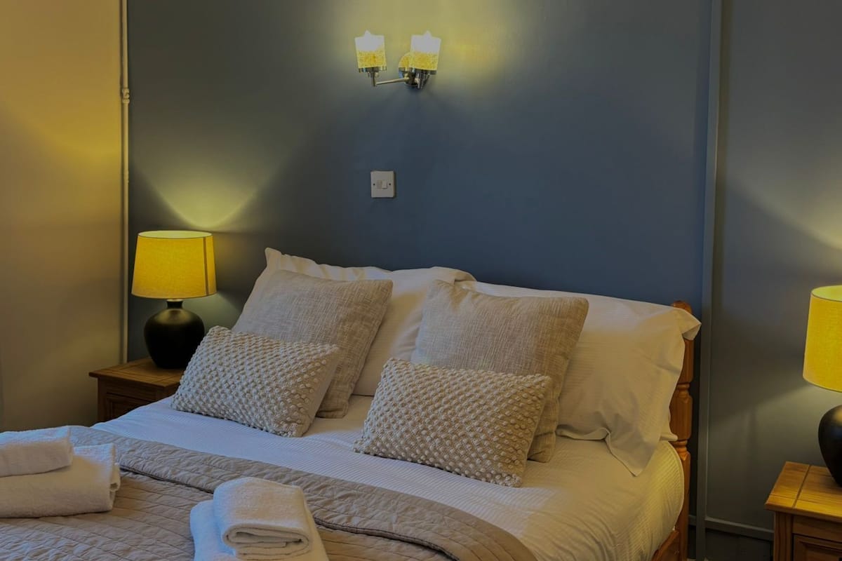 A neatly made bed with a beige quilt, two pillows, and a folded towel on a dark blue wall. There are two bedside lamps with yellow shades, and a light switch on the wall.