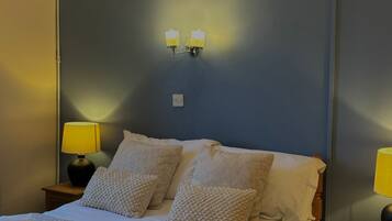 Una cama bien hecha con una colcha color crema, dos almohadas y una toalla doblada sobre una pared azul oscuro. Hay dos lámparas de noche con pantallas amarillas y un interruptor de luz en la pared.