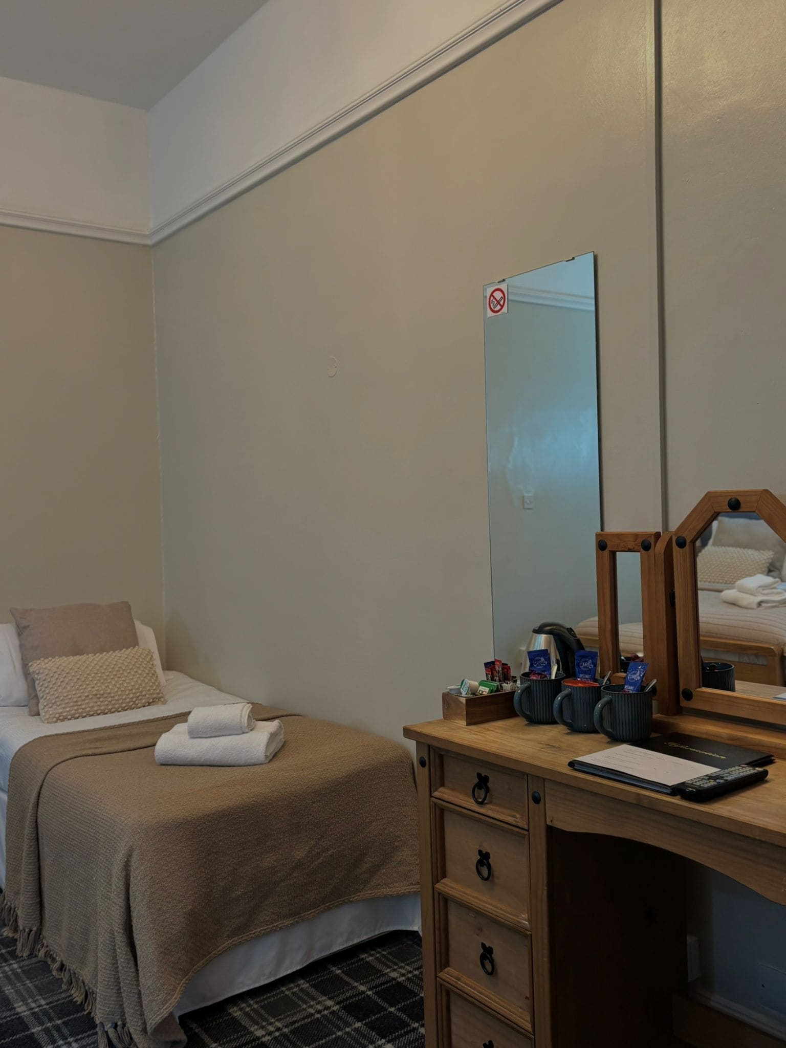 Una habitación de hotel con una cama, un escritorio de madera con espejo, una alfombra a cuadros blanco y negro, y un pequeño armario con cajones.