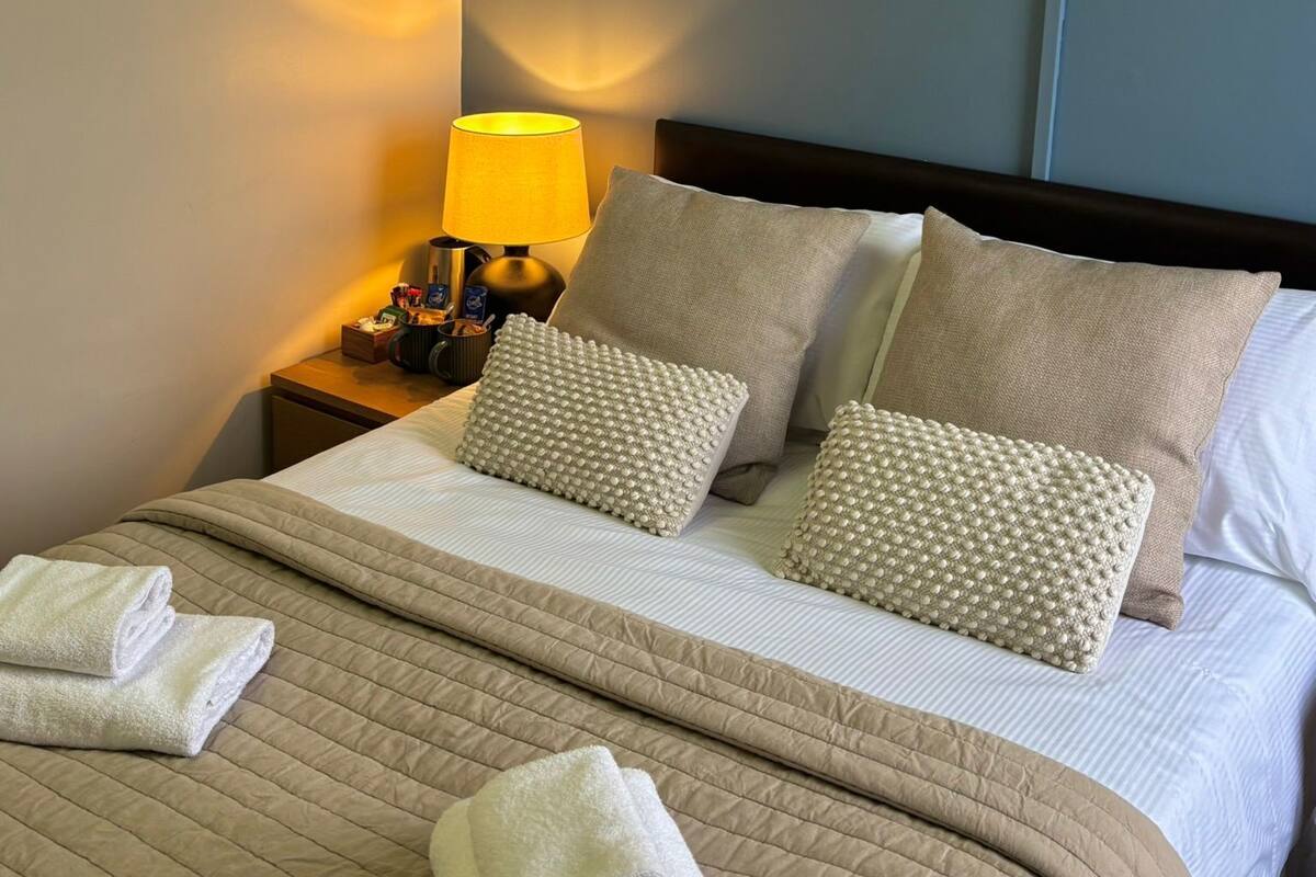 A neatly made bed with two pillows, a bedside lamp, and folded towels on a wooden nightstand.