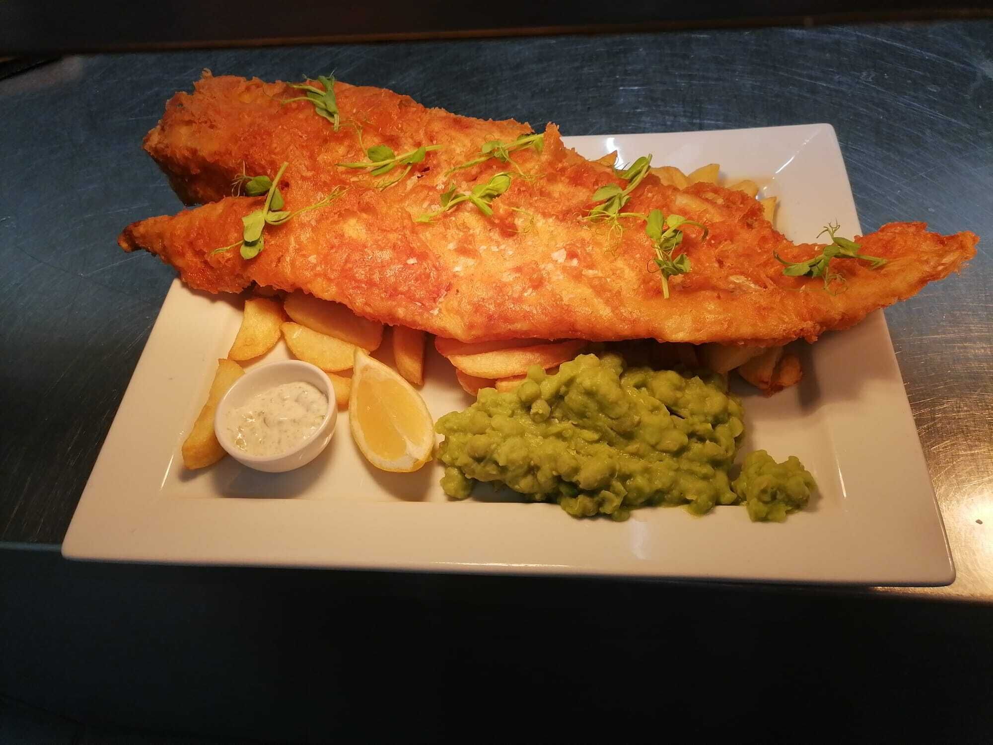 Une assiette de poisson-frites, accompagnée de quartiers de citron, de purée de petits pois et de sauce tartare.