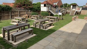 Ein Garten mit Picknicktischen, einer Holzterrasse und einem Haus mit rotem Dach.
