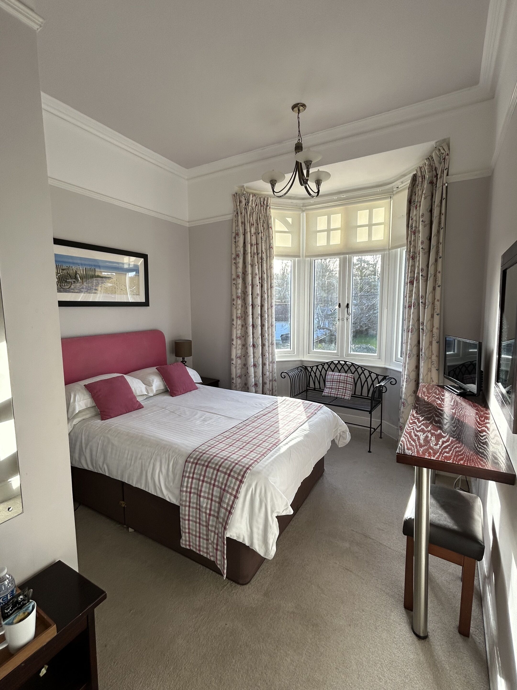 A bedroom with a bed, a desk, a chair, a window with curtains, a chandelier, and a painting on the wall.