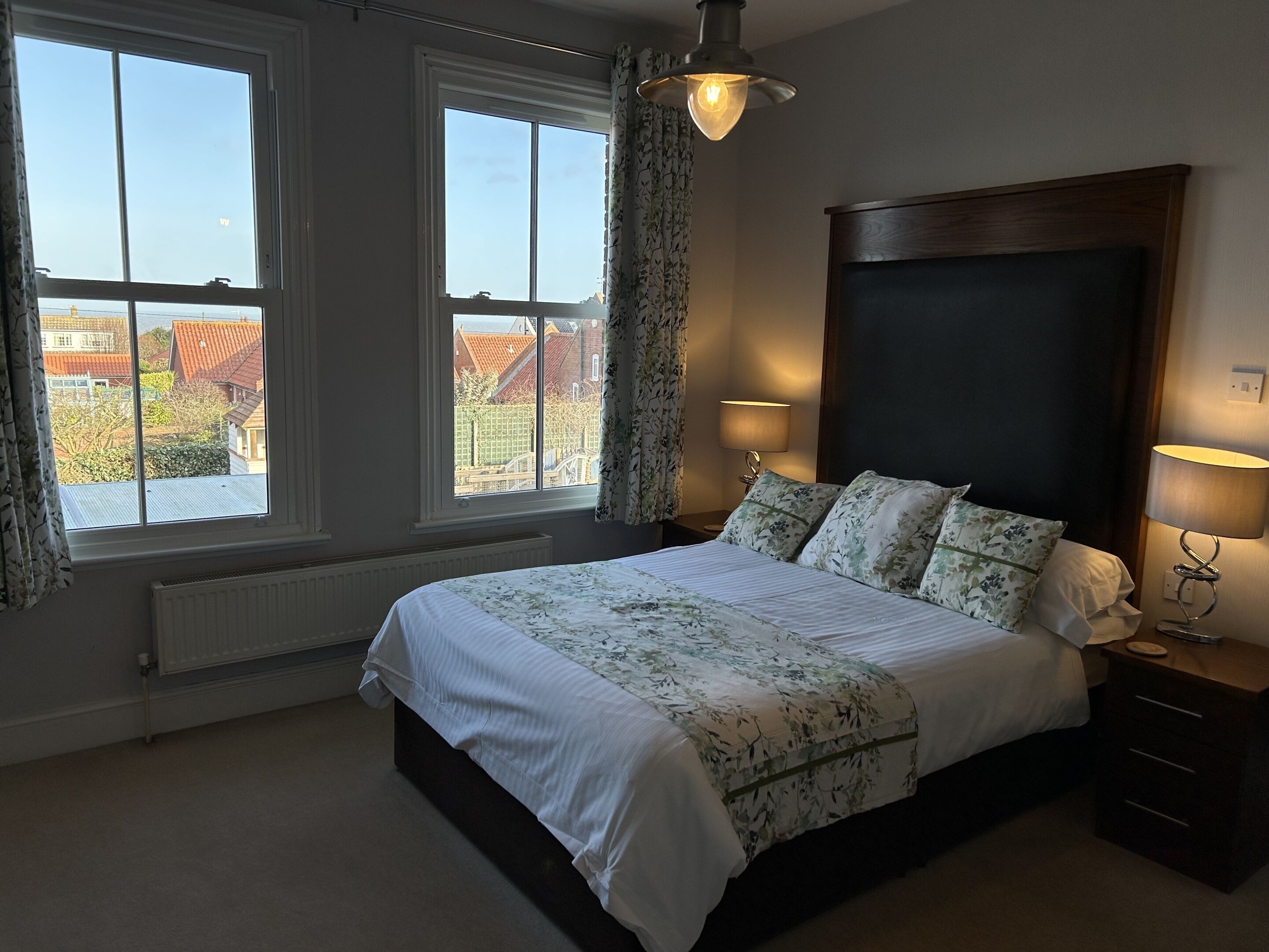A bedroom with a large bed, two bedside lamps, a window with curtains, and a view of buildings outside.