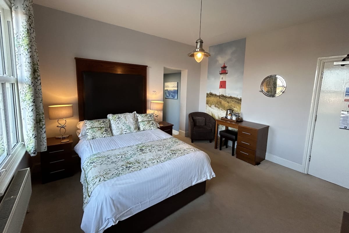 A bedroom with a bed, bedside lamps, a desk, a chair, a painting, and a mirror on the wall.