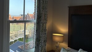 Ein Hotelzimmer mit großem Fenster, einem Bett mit floralem Bettzeug, einem Nachttisch mit Lampe und Blick auf Gebäude und eine Brücke.