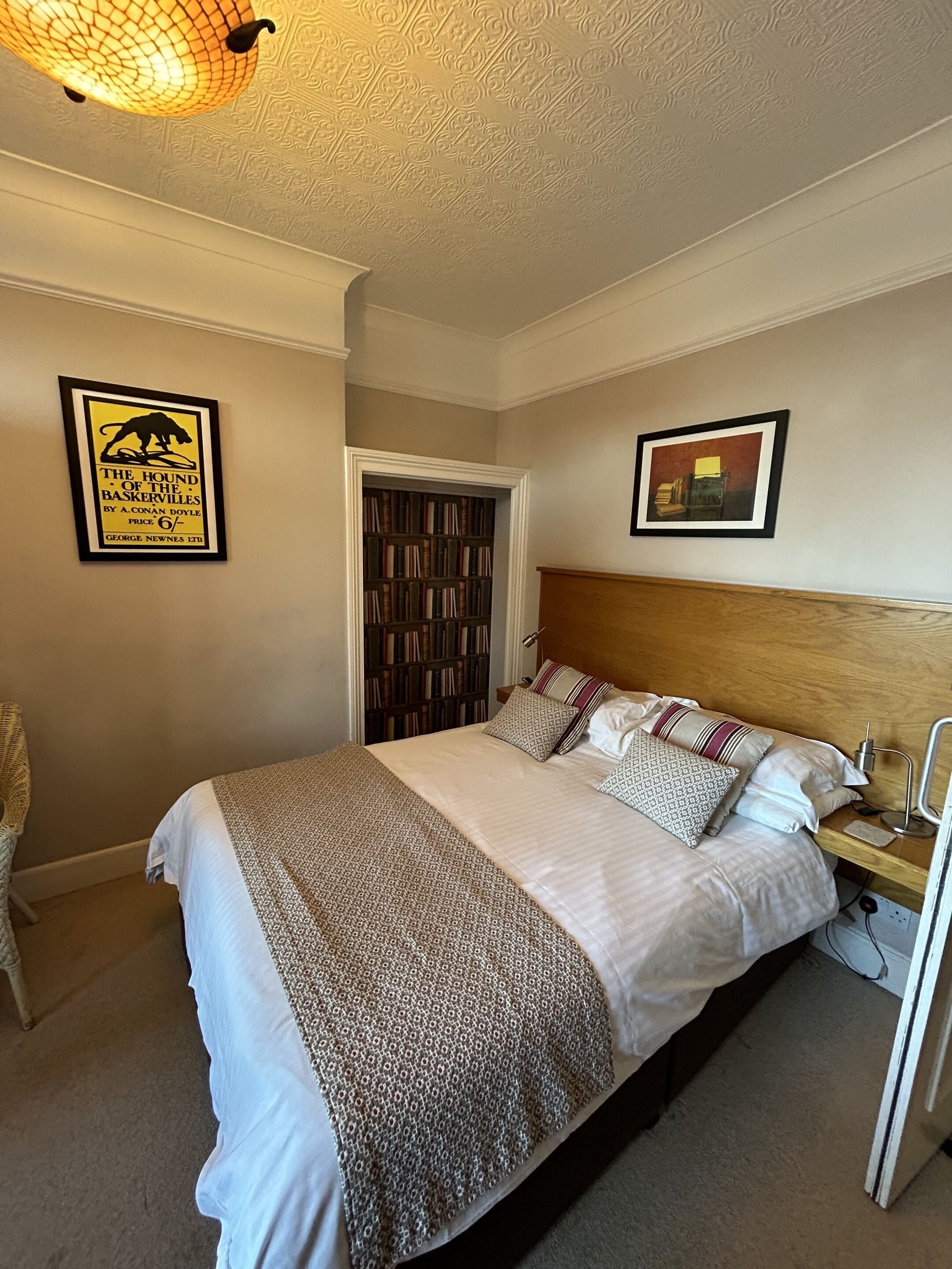 A neatly made bed with patterned bedding, a bedside table with a lamp, a framed book on the wall, and a bookshelf in the corner.