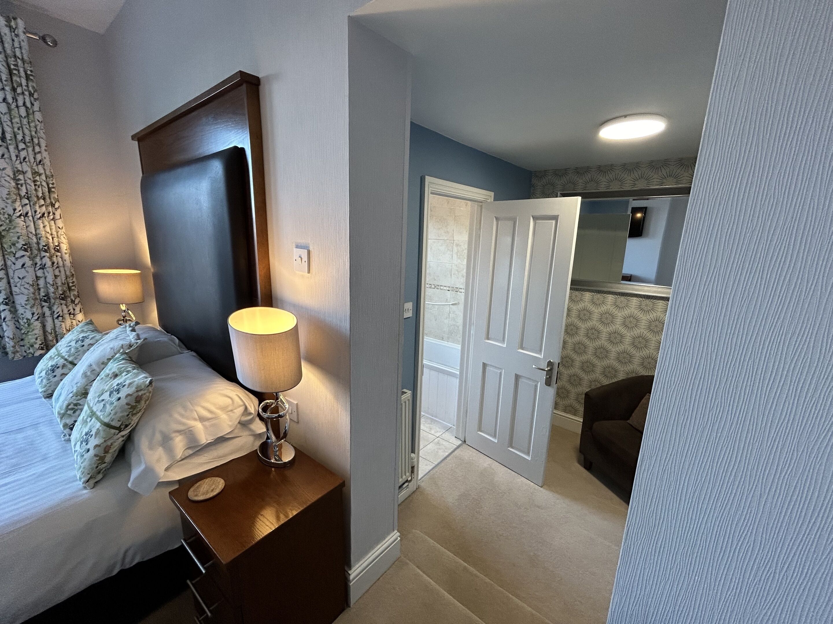 A hotel room with a bed, a bedside table with a lamp, a door leading to a bathroom, and a small seating area.