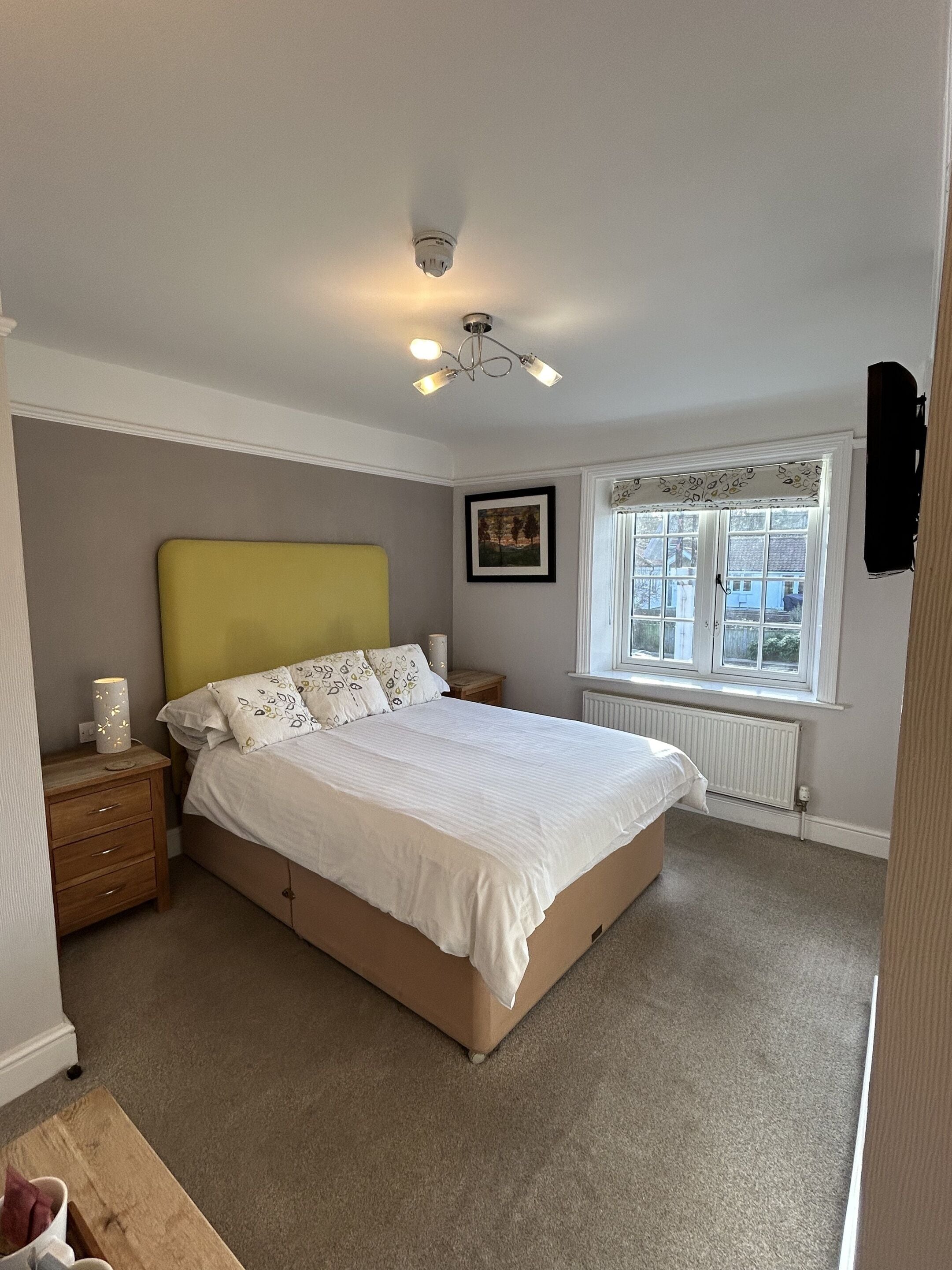 Ein Schlafzimmer mit Bett, Nachttisch, Lampe, Fenster und Heizkörper.