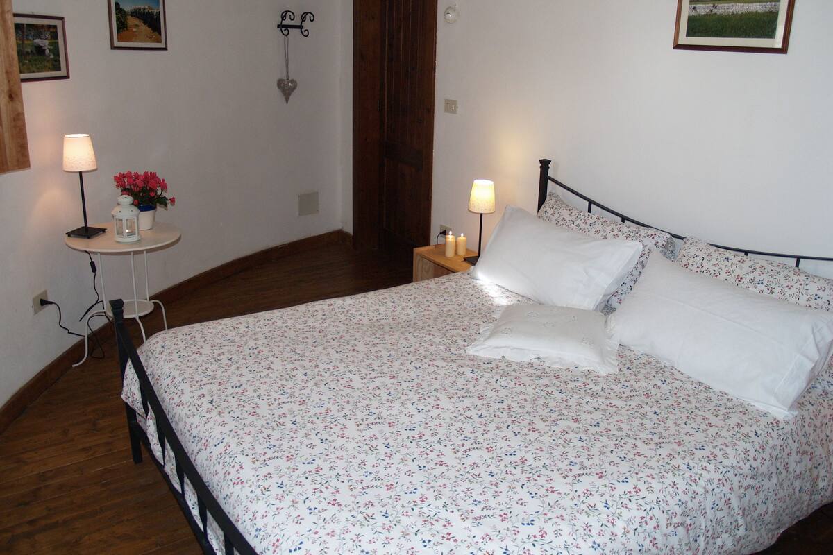 A bedroom with a floral bedspread, a bedside table with a lamp, a small table with flowers, and a picture on the wall.