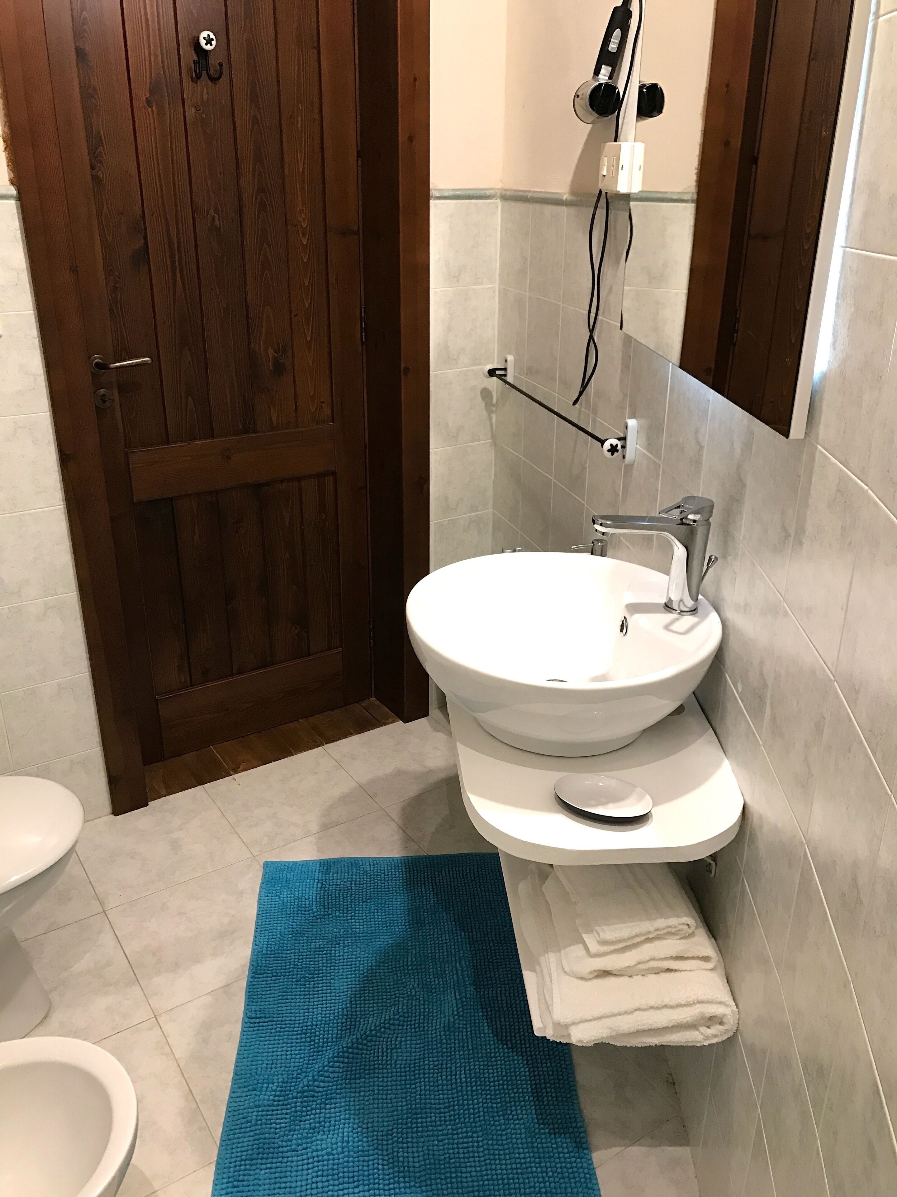 Un baño con puerta de madera, lavamanos blanco, toallero y felpudo azul.
