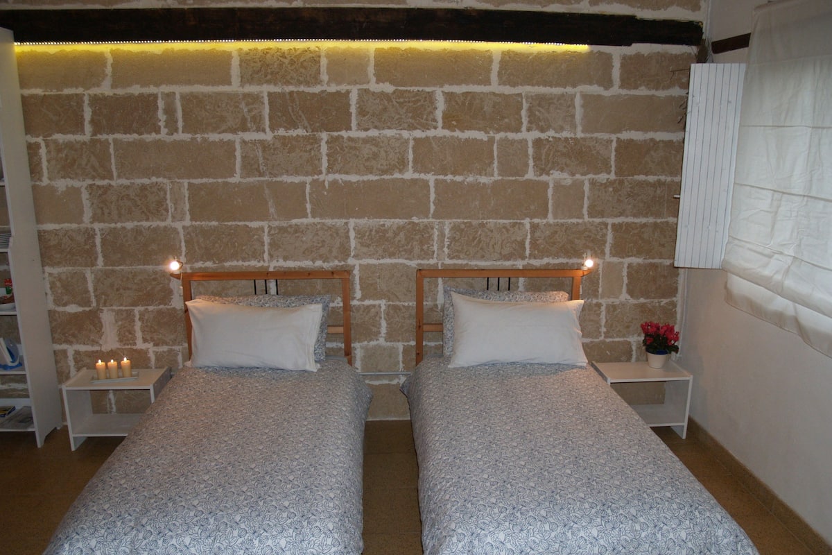 A room with two single beds, a stone wall, a wooden headboard, a nightstand with a lamp, and a small table with a flower.