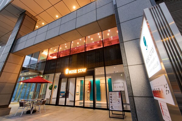 A hotel entrance with a modern design, featuring a glass facade, a red umbrella outside, and a sign indicating the hotel name.