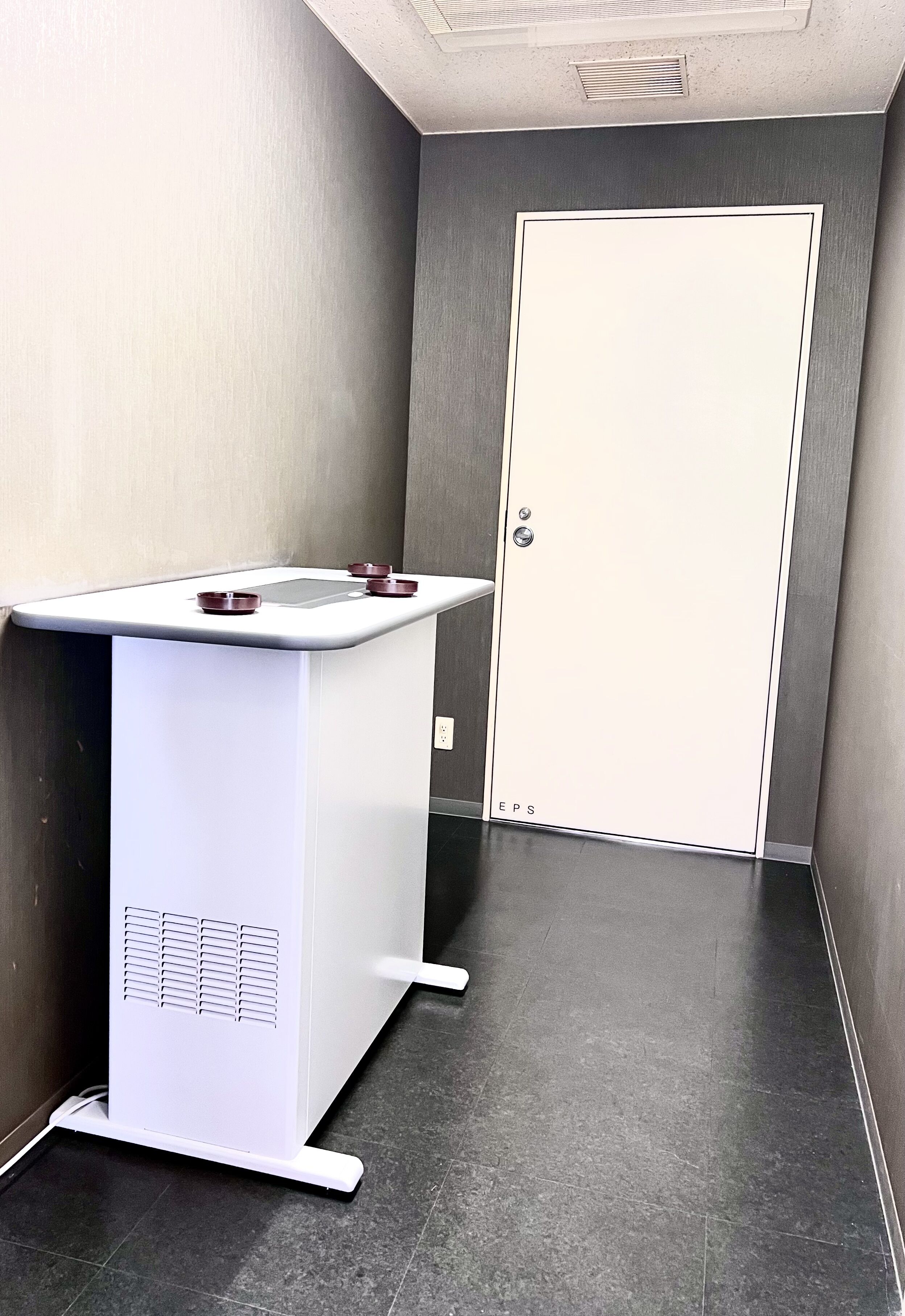 Une petite pièce rectangulaire, blanche, avec une porte, un comptoir blanc doté d’un évier et un sol carrelé.