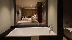 A modern bathroom with a large bathtub, a separate sink, and a mirror reflecting a bedroom with a bed and curtains. - The Swiss Residence (Kandy)