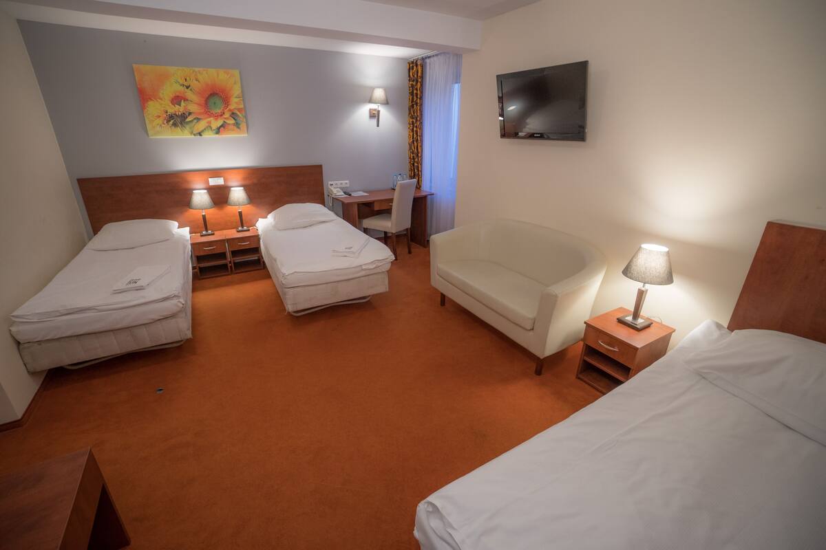 A hotel room with two single beds, a desk with a chair, a sofa, a TV mounted on the wall, and a painting above the desk.