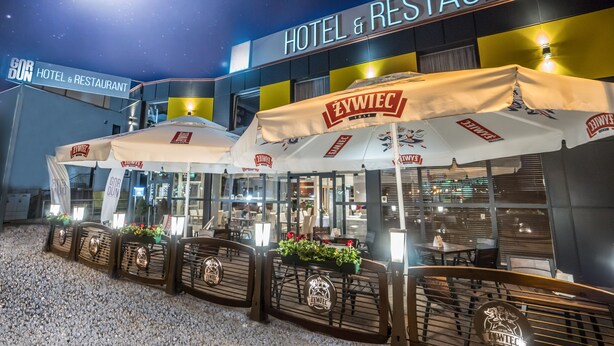 An outdoor seating area of a hotel and restaurant with branded umbrellas and a clear view of the interior. - Gordon Hotel (Warsaw)