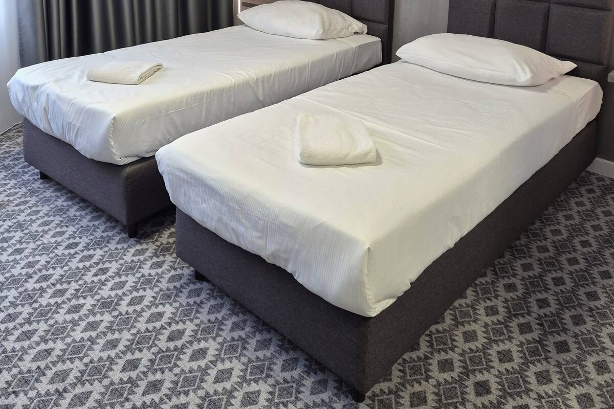 A hotel room with two beds, a grey headboard, a patterned carpet, and a window with curtains.