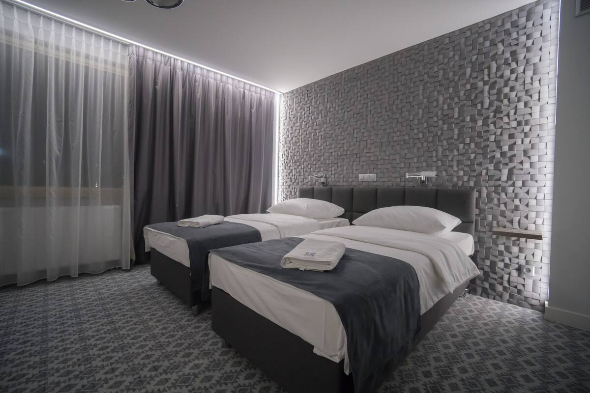 A hotel room with two beds, a large window with sheer curtains, a textured wall, and a small shelf with a lamp.