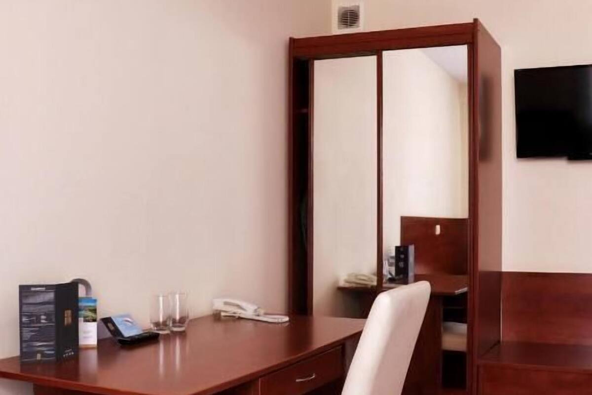 A hotel room with a bed, a desk with a chair, a mirror, a television, and a red carpet.