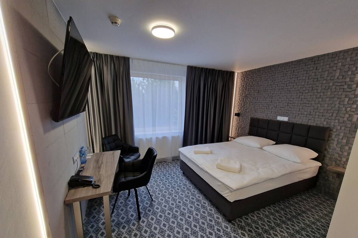 A hotel room with a bed, a desk with a chair, a television, and a window with curtains.