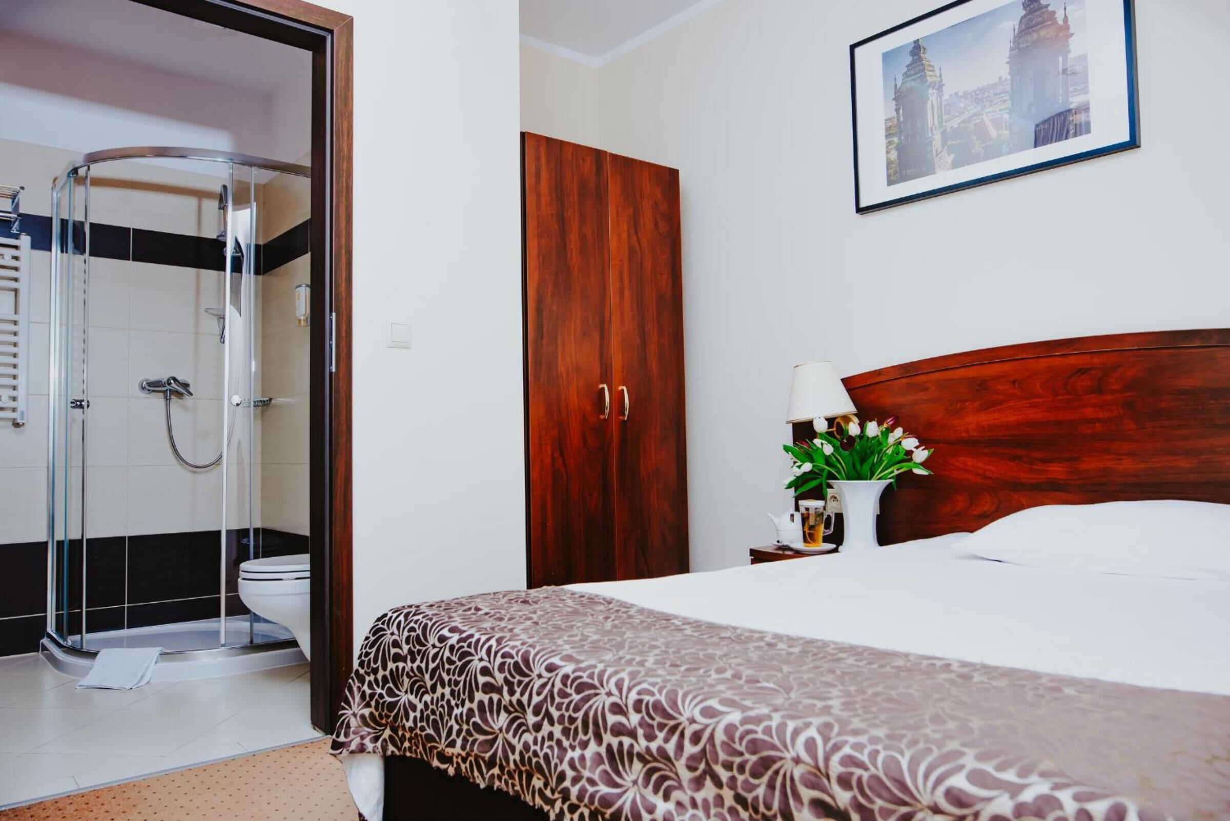 A hotel room with a shower, a wooden wardrobe, a bed with a patterned bedspread, a nightstand with a vase of flowers, and a framed picture on the wall.