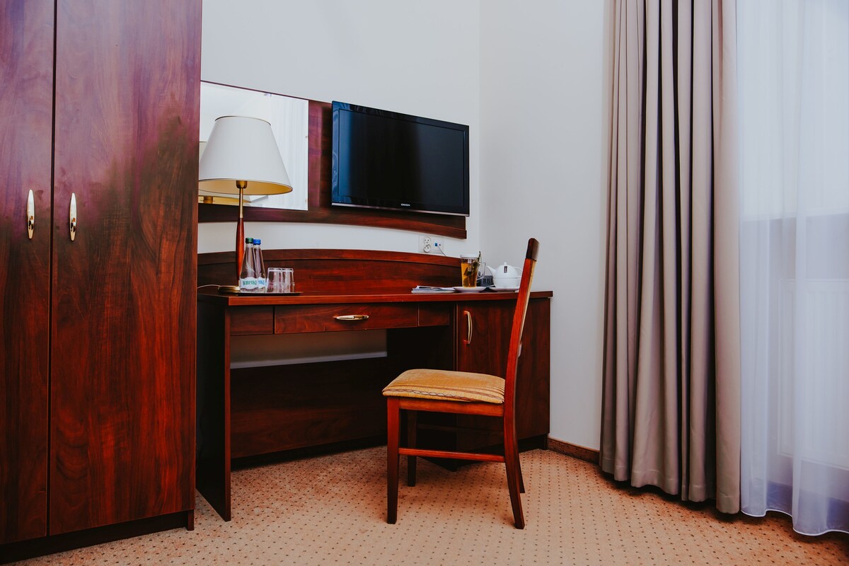 A hotel room with a wooden desk, a television, a lamp, a chair, and a window with curtains.