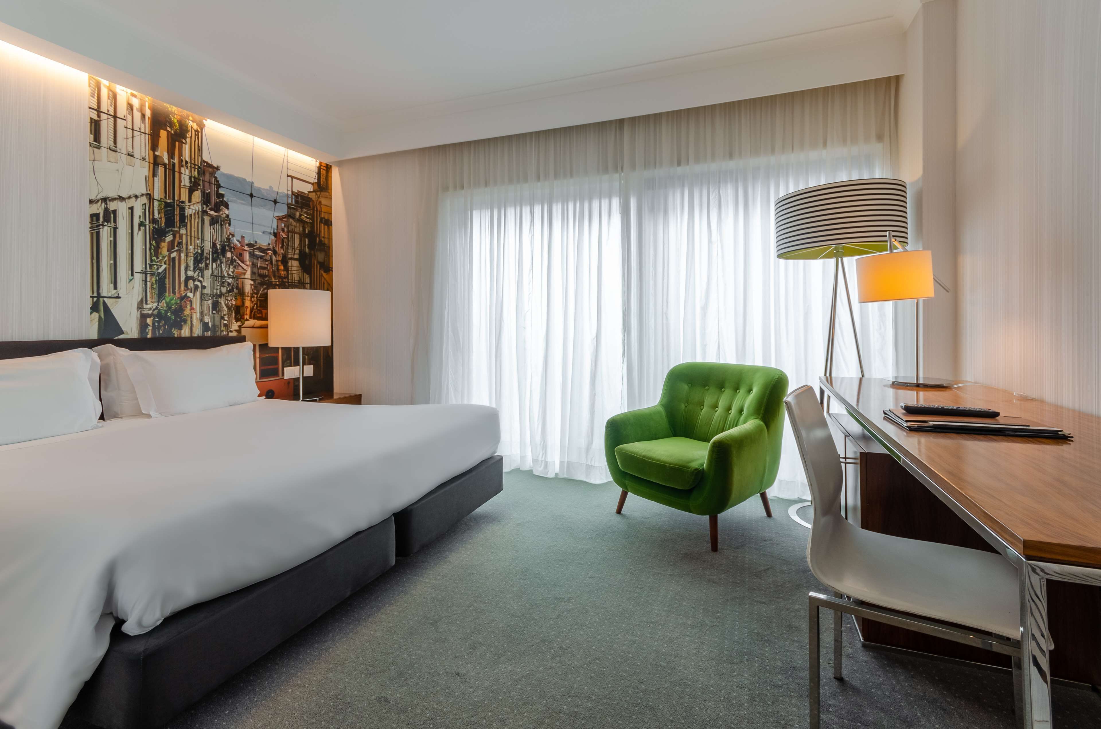 A hotel room with a bed, a desk, a green armchair, a lamp, and a large window with sheer curtains.