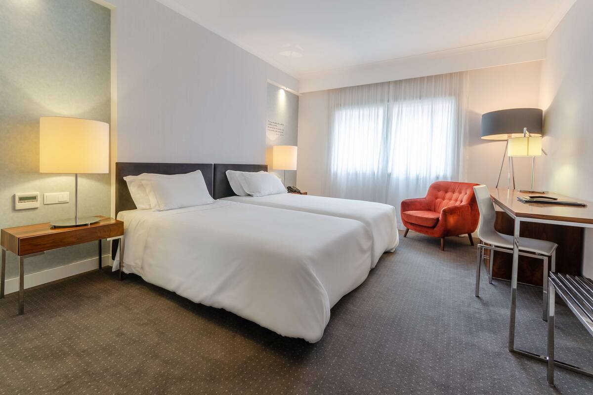 Une chambre d’hôtel avec deux lits, un bureau avec une chaise, une lampe et une fenêtre avec des rideaux.