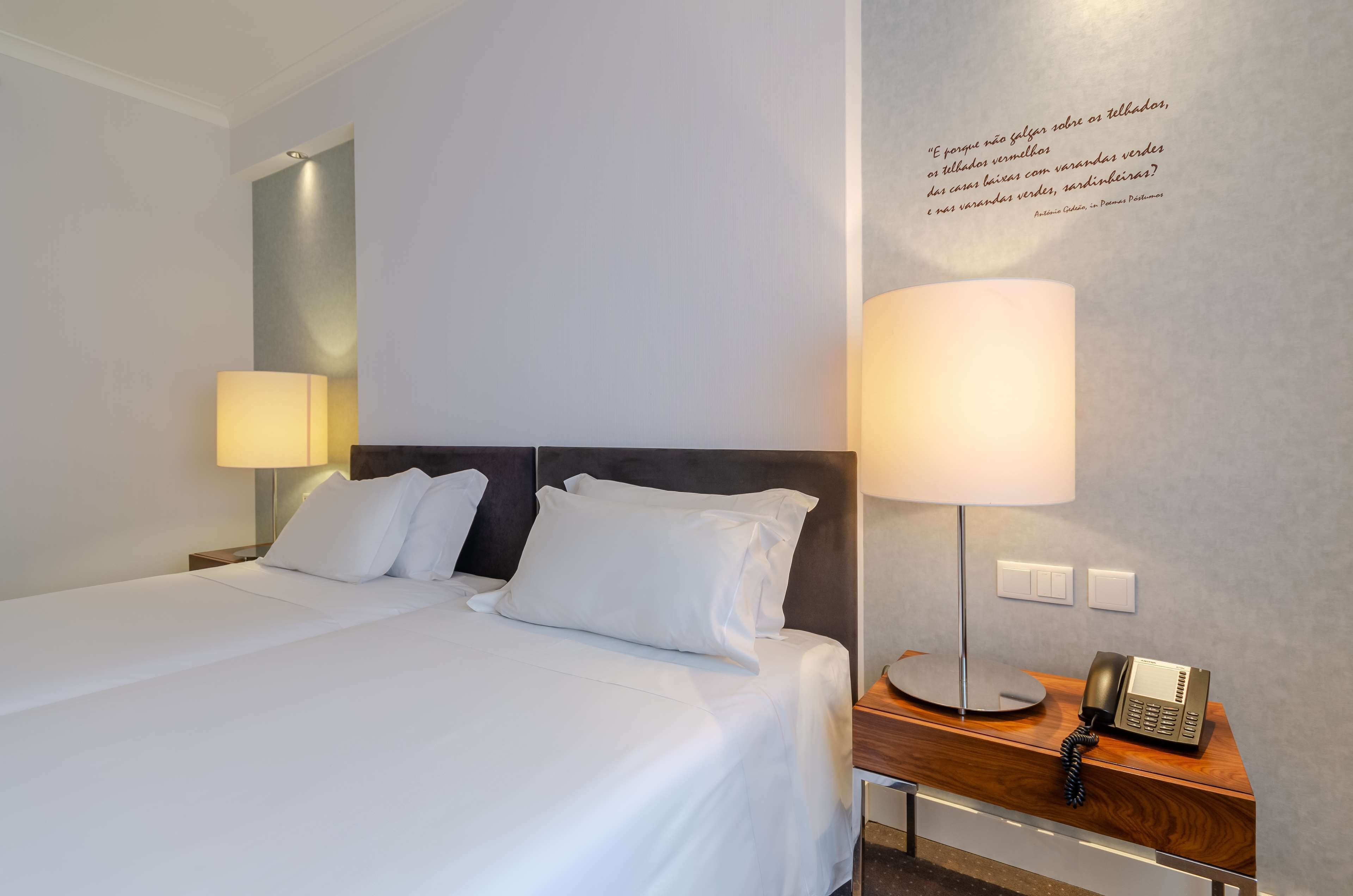 Ein Hotelzimmer mit einem ordentlich bezogenen Bett, einer Nachttischlampe, einem Telefon und einer Wand mit einem Zitat.