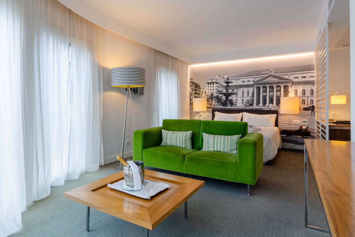 Une chambre d’hôtel moderne avec un canapé vert, une table basse en bois et un lit doté d’une tête de lit blanche.