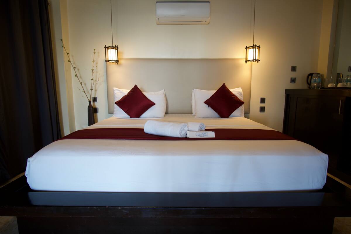 Una habitación de hotel con una cama grande, dos lámparas de noche, un aire acondicionado de pared y un mueble pequeño.