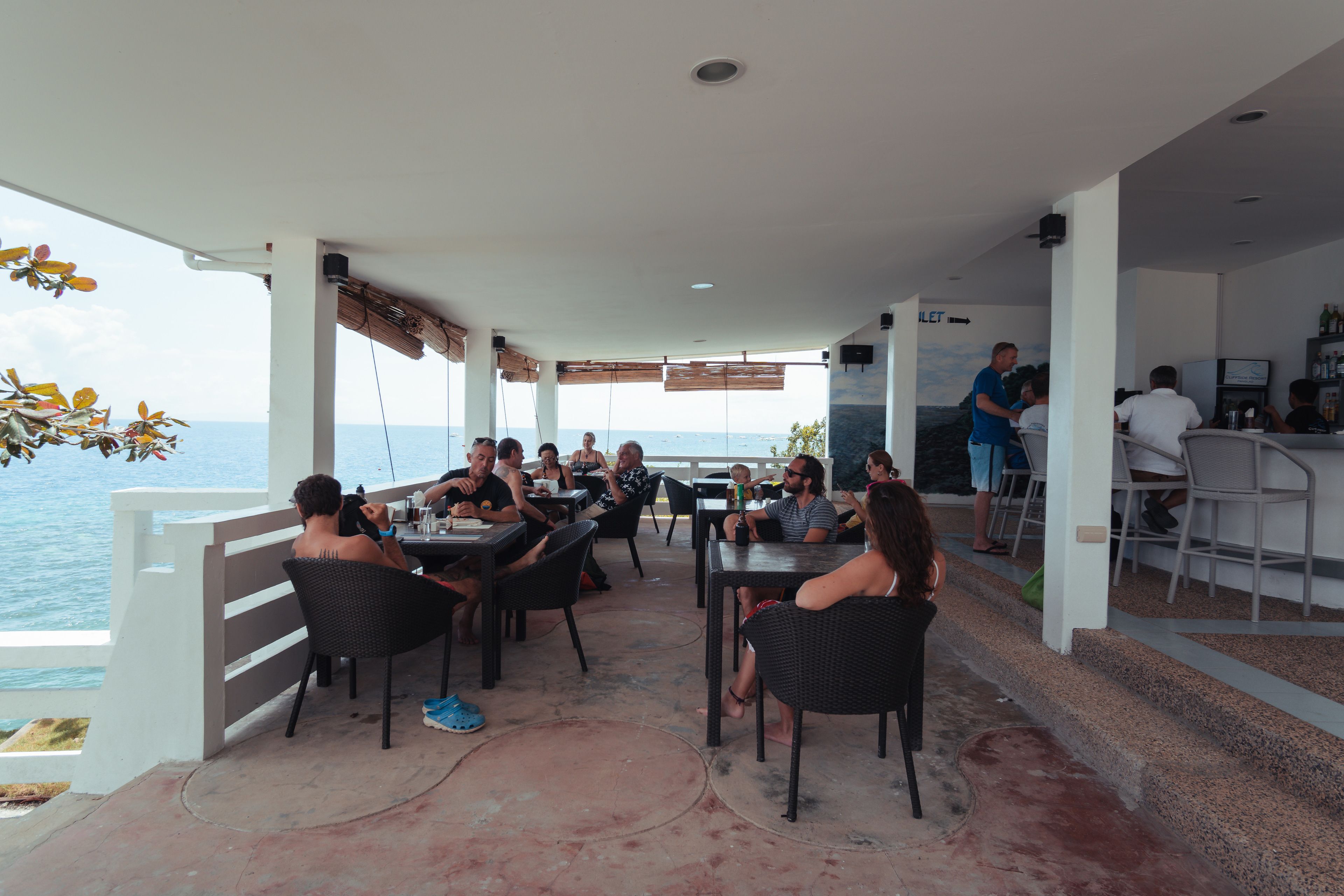 A restaurant with outdoor seating overlooking the sea, featuring a bar area and a view of the ocean.