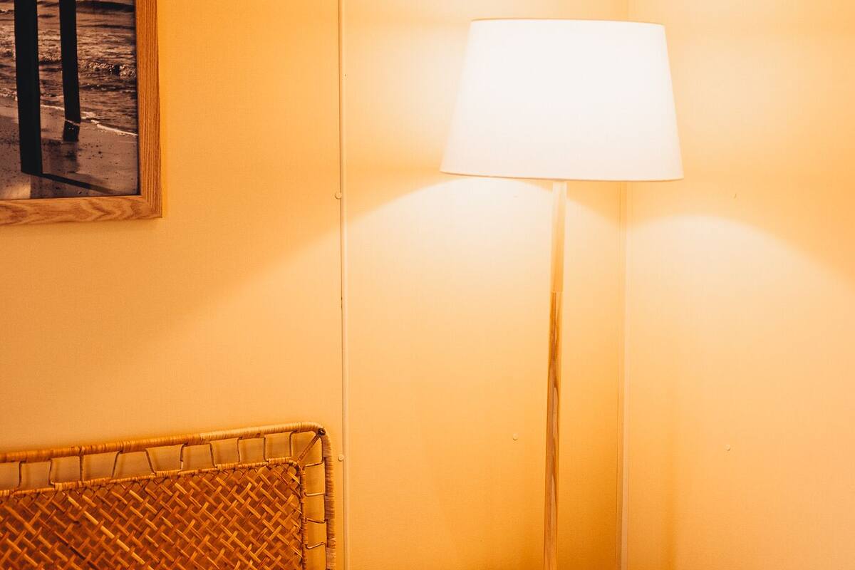 A bed with white linens, a wicker headboard, a standing lamp with a white shade, and a framed picture on the wall.