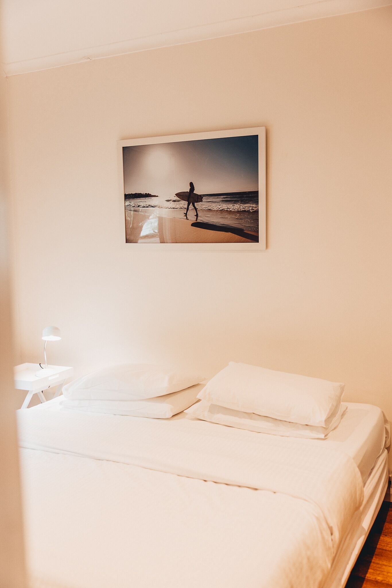 A neatly made bed with white linens in a room with a single wall-mounted picture of a beach scene.