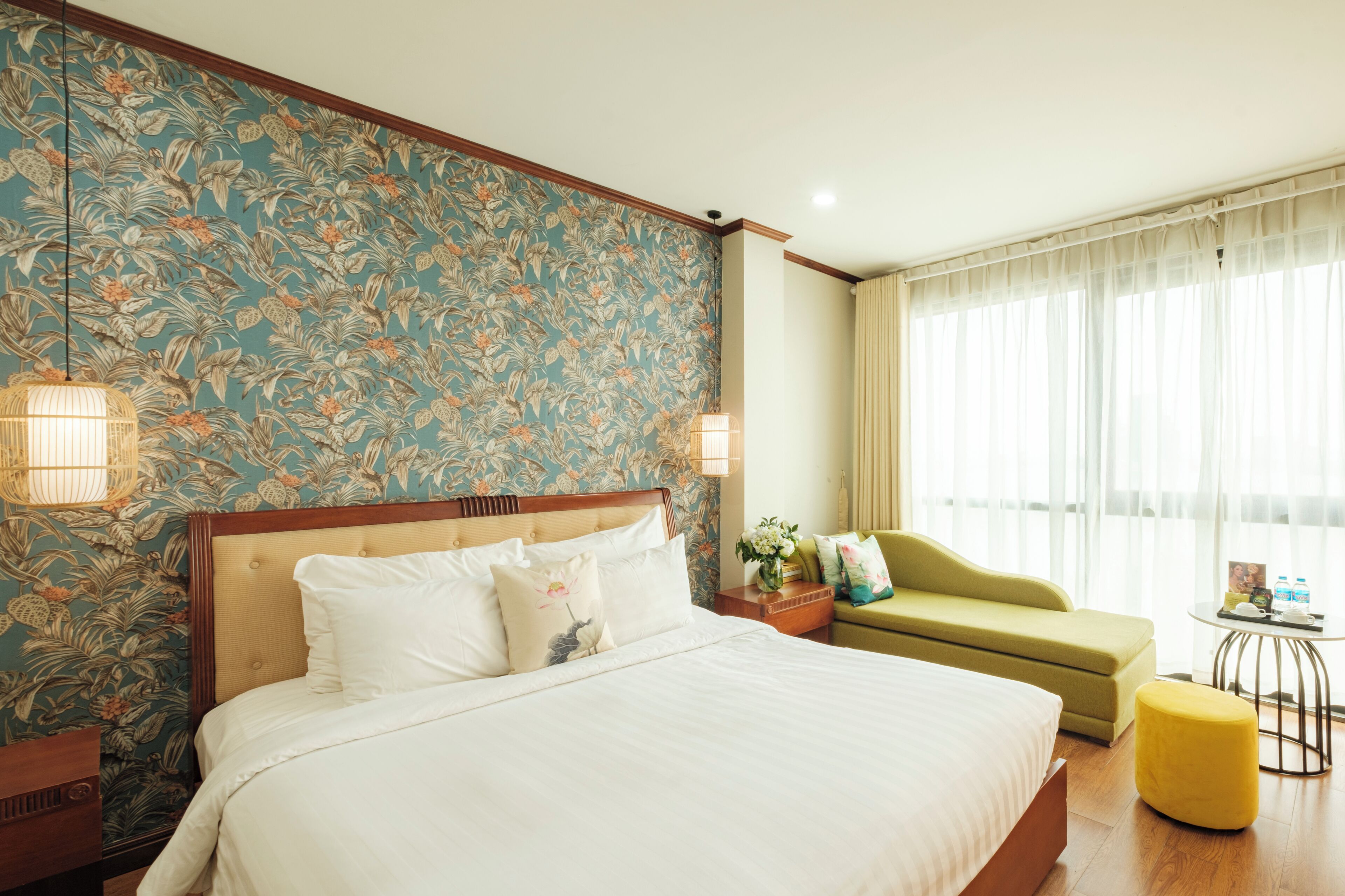 Une chambre d’hôtel avec un grand lit, un fauteuil vert, une petite table ronde et une fenêtre avec des rideaux légers.