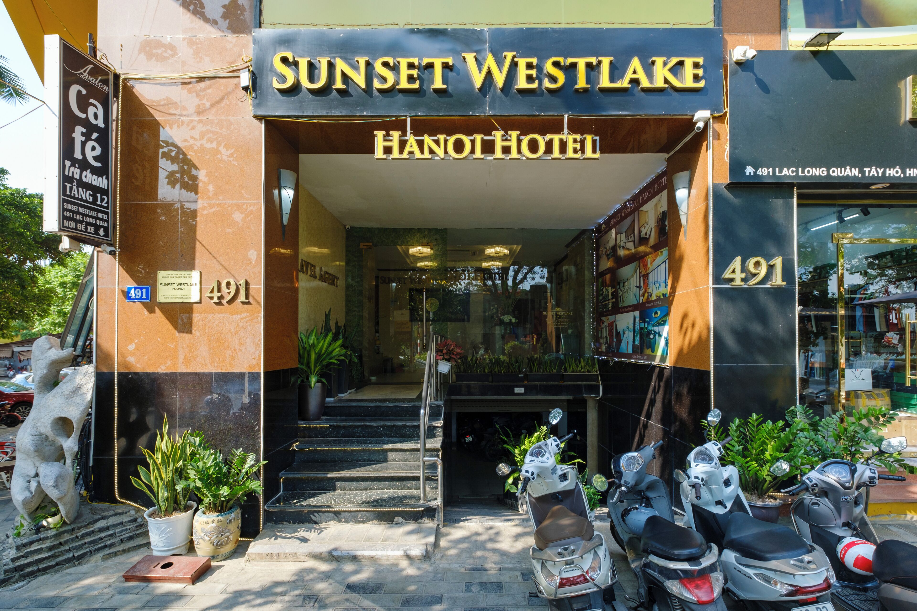 The entrance to the Sunset Westlake Hanoi Hotel, featuring a prominent address sign, a staircase leading to the entrance, and several potted plants.