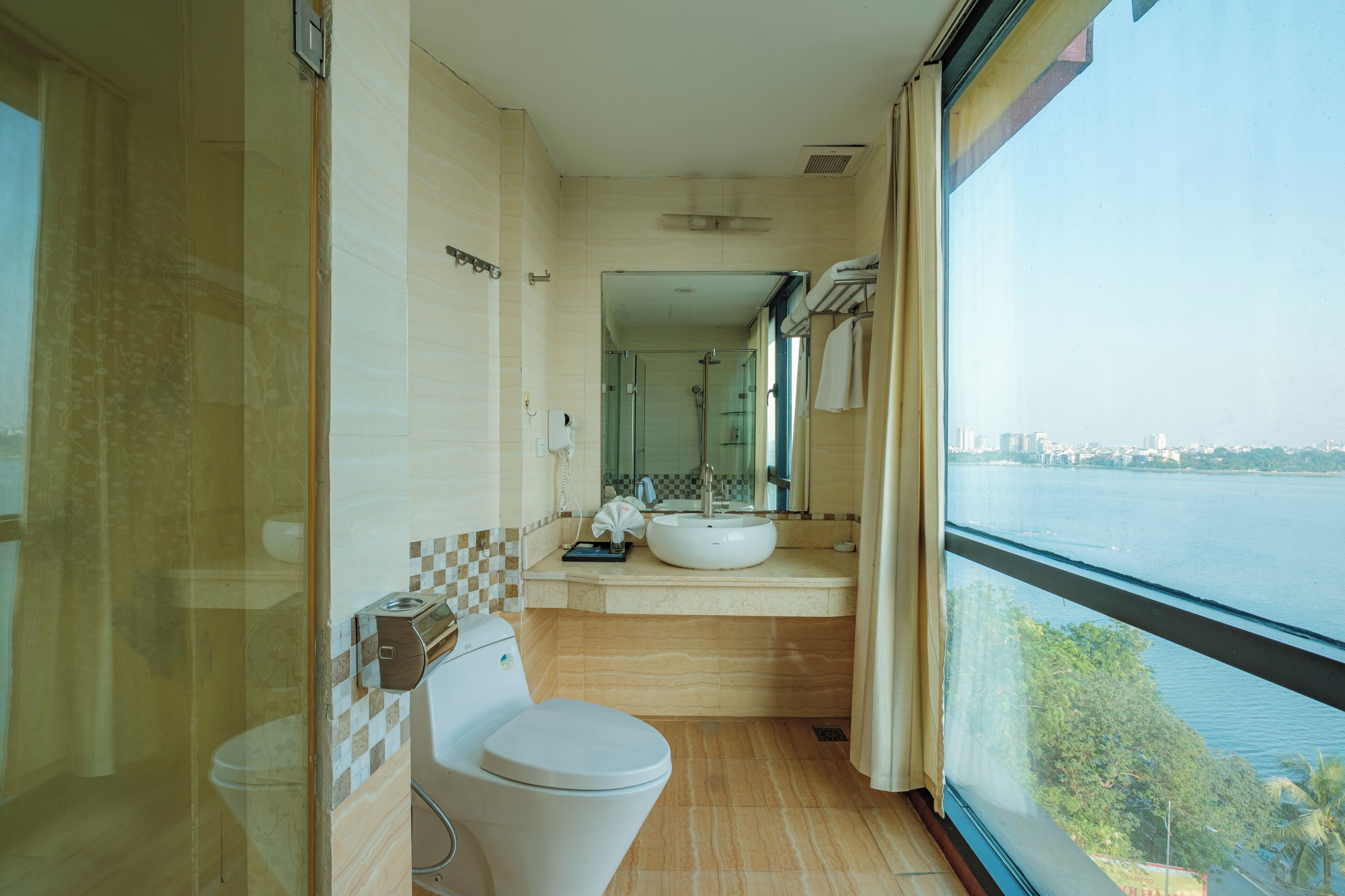 Un baño moderno con inodoro, lavamanos y cabina de ducha de vidrio. Cuenta con un ventanal con vistas a un cuerpo de agua y edificios.