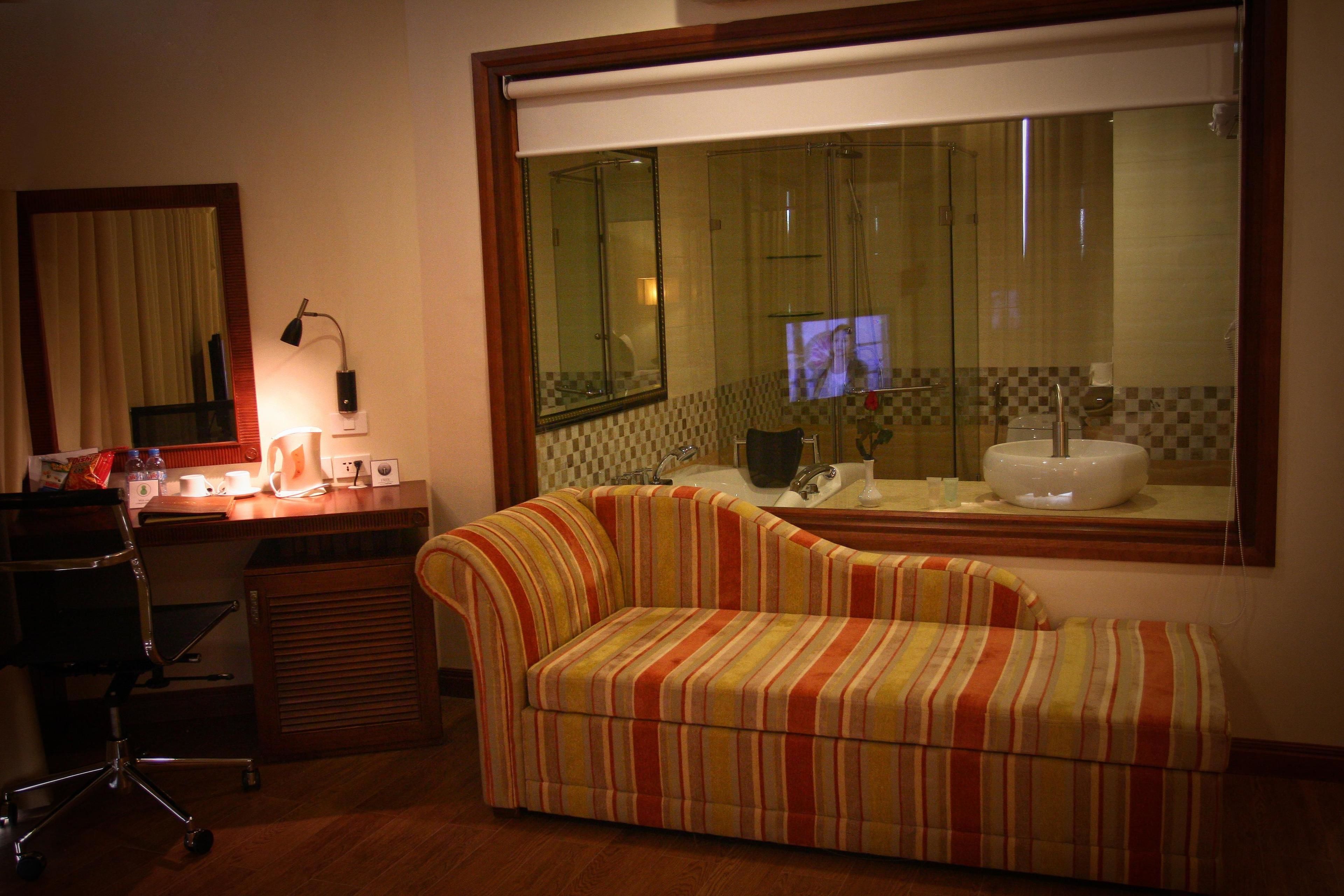 A room with a striped sofa, a desk with a lamp, a chair, and a large window with a view of a bathroom.