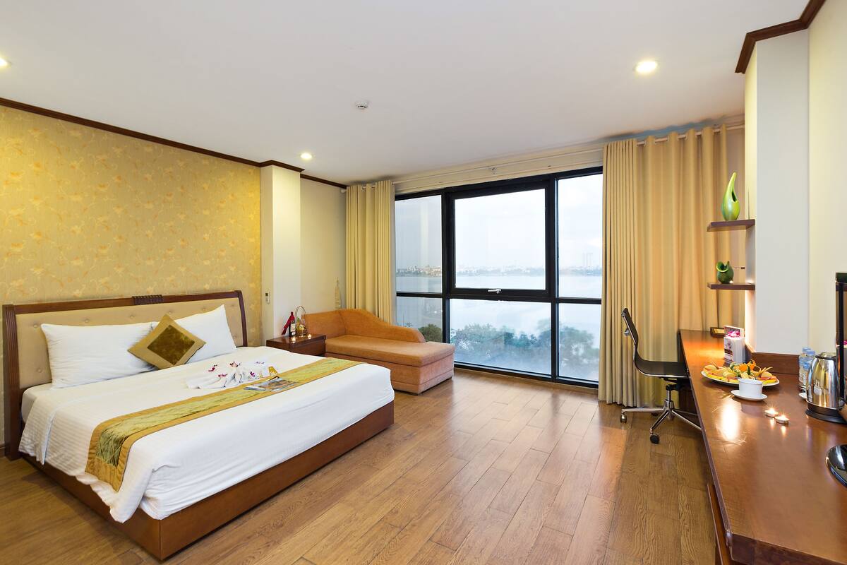Habitación de hotel con una cama grande, un sofá, un escritorio con una silla, vistas de la ciudad y un ventanal con cortinas.