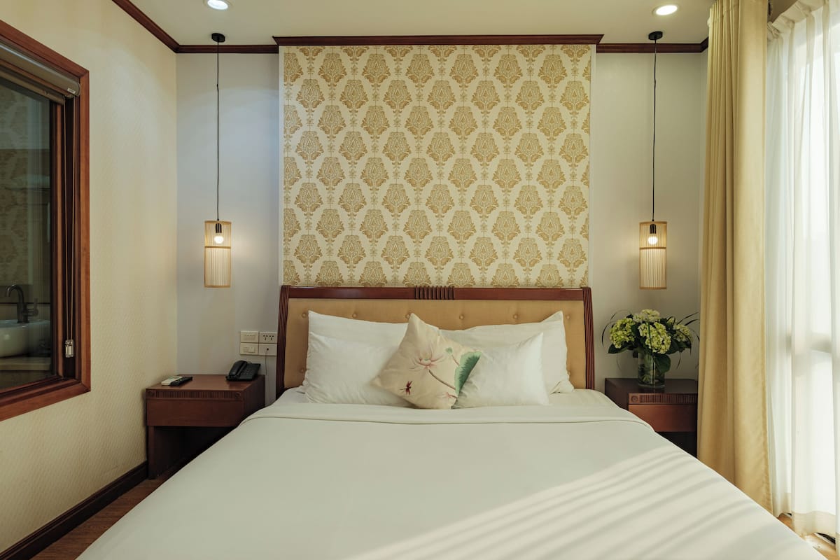Una cama bien tendida con sábanas blancas y almohadones decorativos, un cabecero de madera, mesitas de noche con lámparas y un papel tapiz estampado sobre la cama.