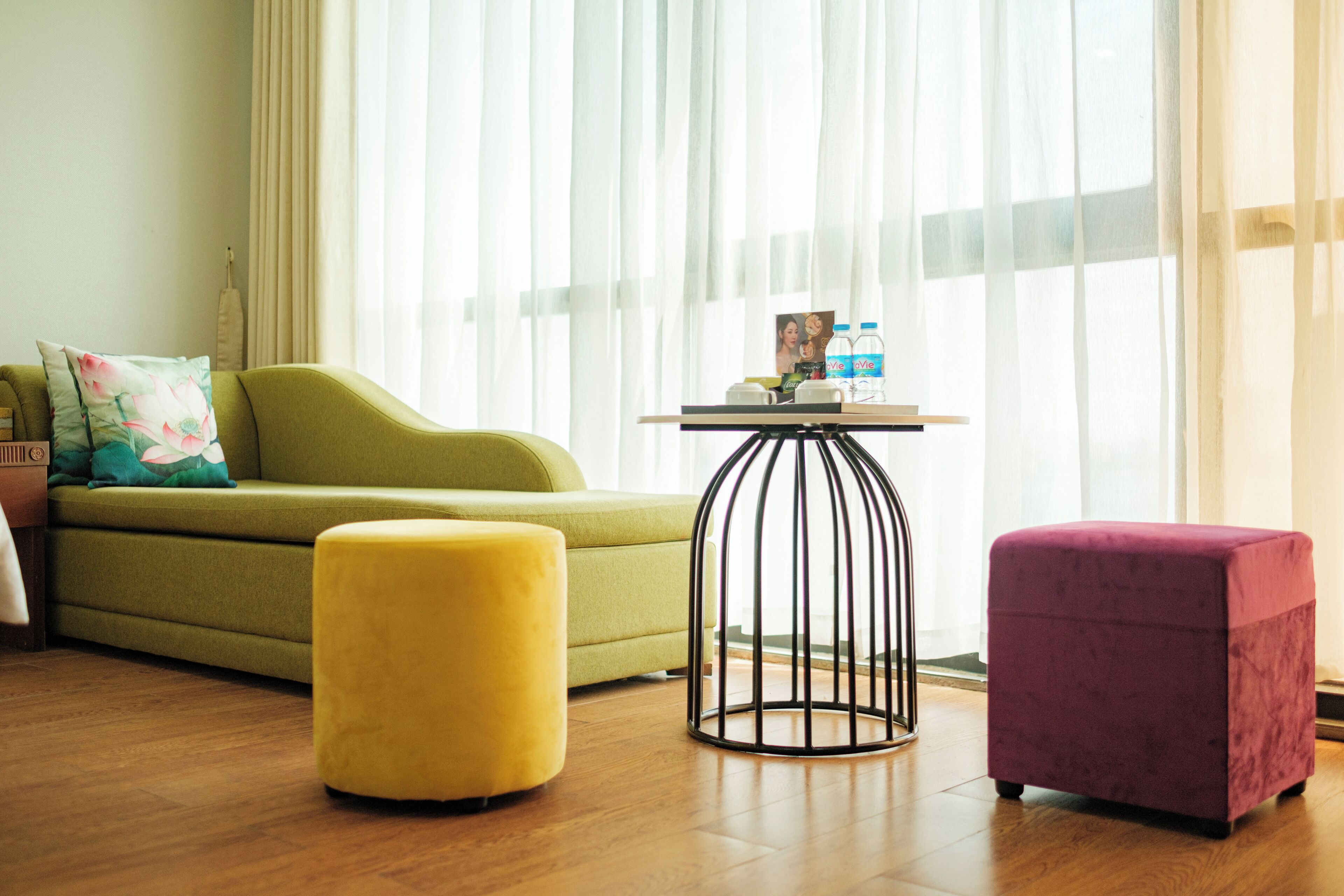 A living room with a green sofa, a yellow ottoman, a purple ottoman, a small round table with a photo and water bottles, and sheer curtains.