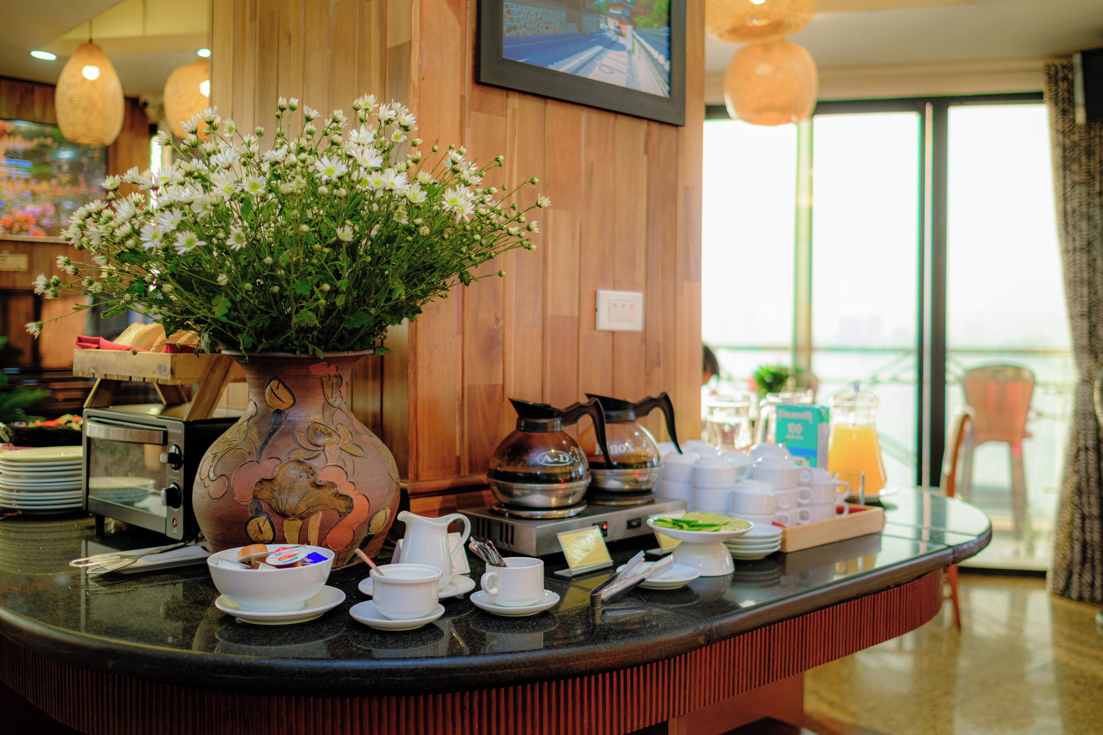 A hotel reception area with a wooden counter, a vase of flowers, a coffee maker, and breakfast items.