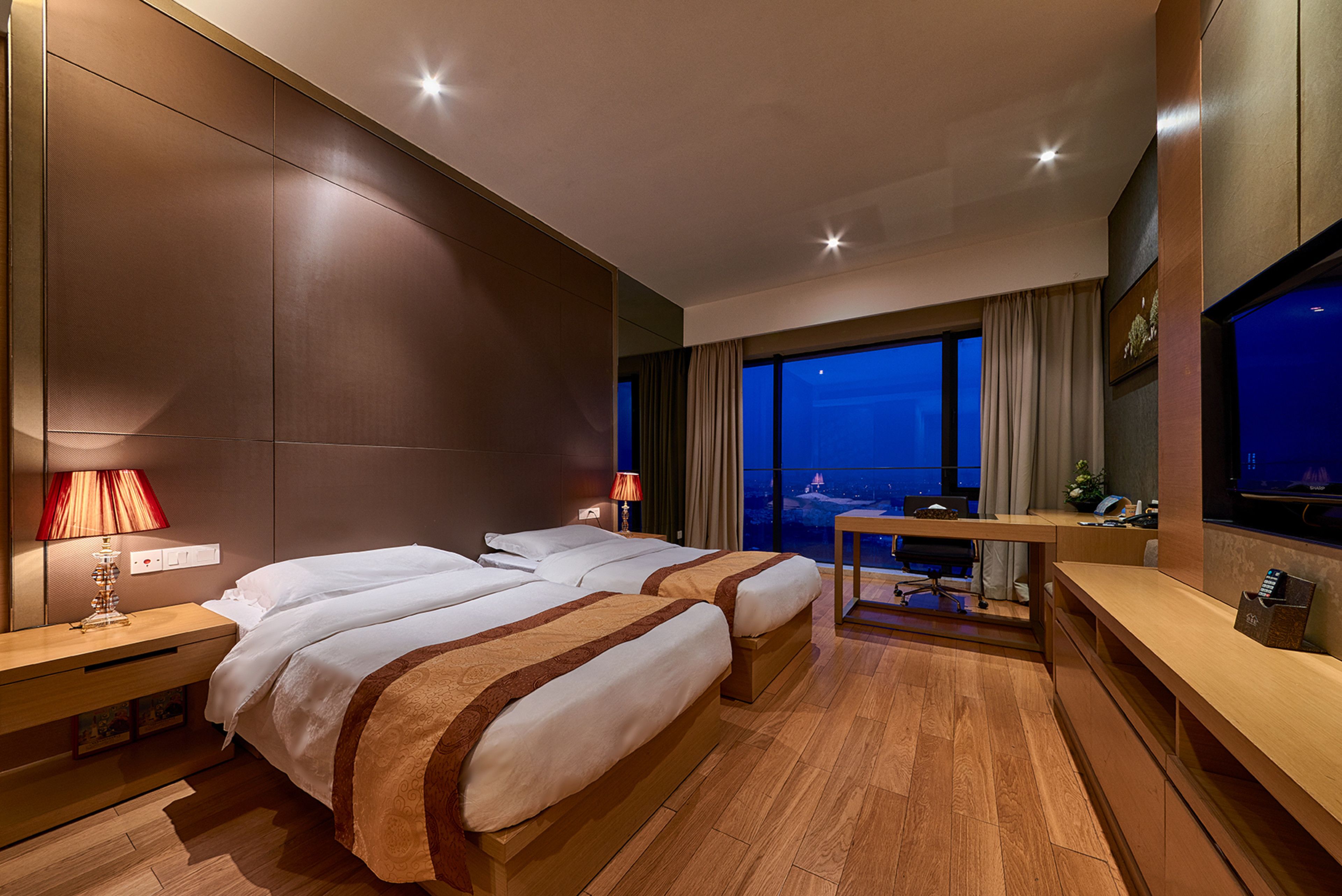 A hotel room with two beds, a desk, a TV, and a view of the city at night.