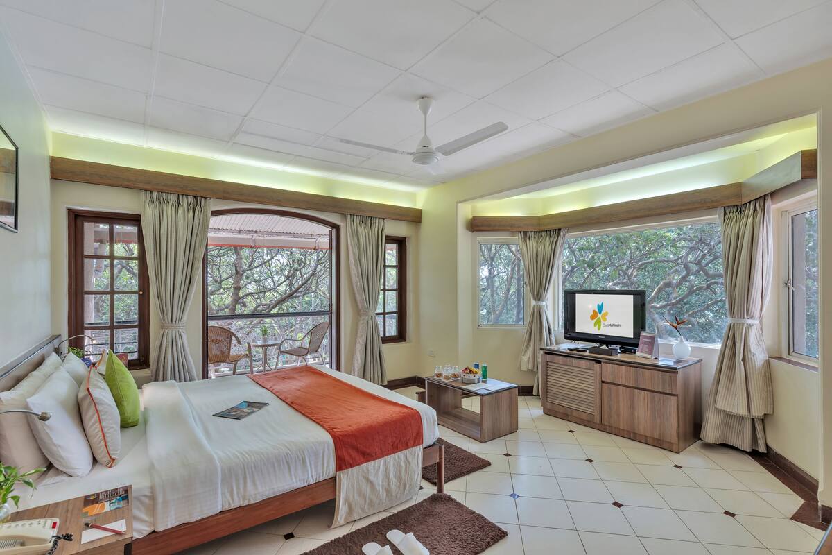 Una habitación de hotel con cama, televisión, escritorio y vistas a árboles a través de amplios ventanales.