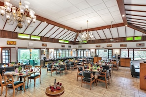 Un restaurante espacioso con techos altos, vigas de madera y ventanas grandes. Las mesas están dispuestas con sillas, y hay varias zonas de comedor.