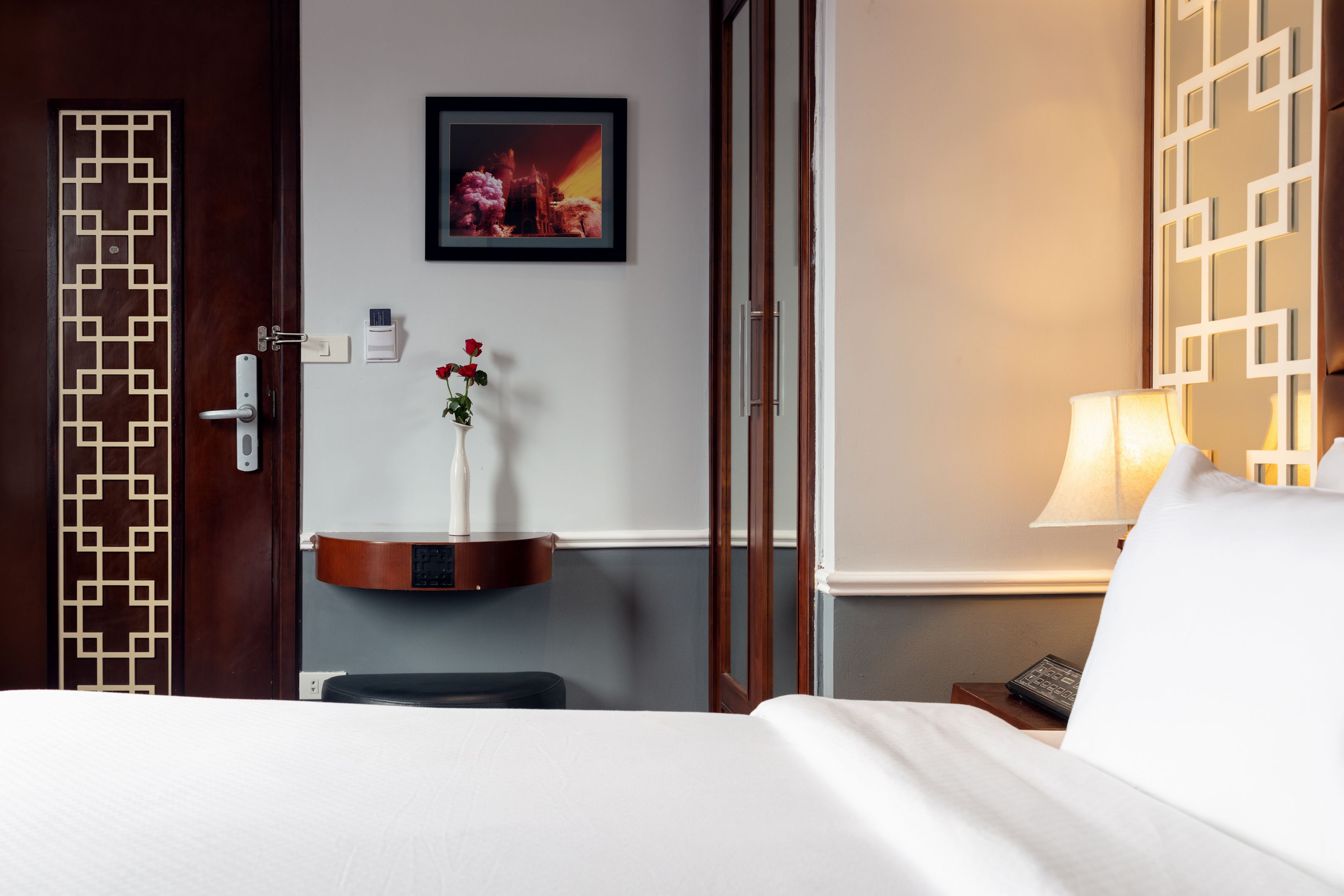 Une chambre d’hôtel avec un lit, une table de chevet avec une lampe, un tableau au mur et un panneau décoratif sur la porte.